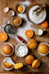Obraz na płótnie Canvas Homemade citrus oranges or clementines sweet muffins cakes with flour and ingredients above over wooden cutting board as background. Flat lay, space