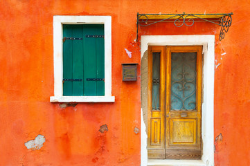 Fototapeta na wymiar Window with shutters and door on the orange facade of the house. Colorful architecture in Burano island, Venice, Italy.