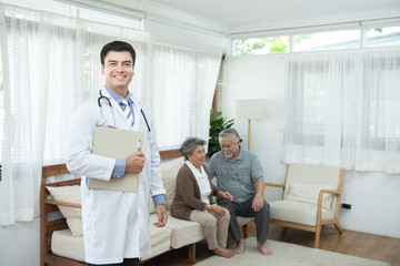 Young handsome caucasian male doctor standing hand holding document file look at camera with smile and two elderly old senior asian couple sit on couch in background,healthcare and medical concept.