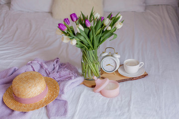 A bouquet of tulips in a glass vase, a white alarm clock, a candle, a note, a cappuccino mug on a wooden board are standing on the bed. Women's things. Breakfast in bed.