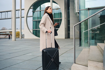 Side view of stylish businesswoman waiting on street with cellphone and suitcase