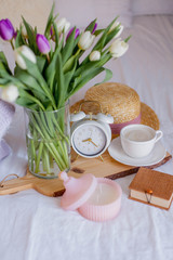 A bouquet of tulips in a glass vase, a white alarm clock, a candle, a note, a cappuccino mug on a wooden board are standing on the bed. Women's things. Breakfast in bed.