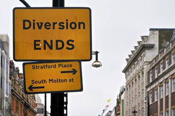 Fototapeta premium Yellow and black Diversion Ends sign with arrows pointing to Stratford Place and South Molton street, blurred building in background