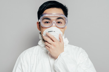 asian epidemiologist in hazmat suit touching respirator mask while looking at camera isolated on grey