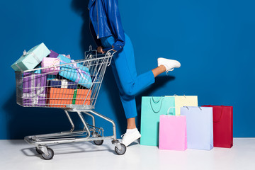 cropped view of african american woman with shopping cart full of gifts near shopping bags on blue...