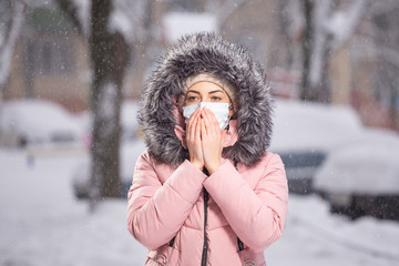 Portrait of woman in pink walking down the street in winter under snowfall in a protective mask to...