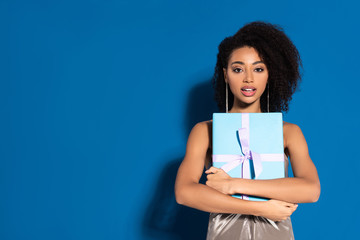 Fototapeta premium excited beautiful african american woman in silver dress with open mouth holding gift on blue background