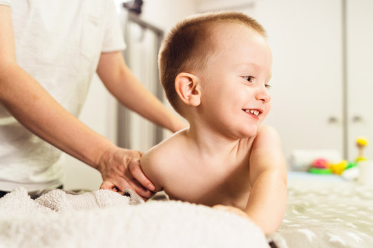 Small caucasian boy laying on the bed naked having back massage by professional female physiotherapist masseuse in a rehabilitation center salon or home office of physical therapy