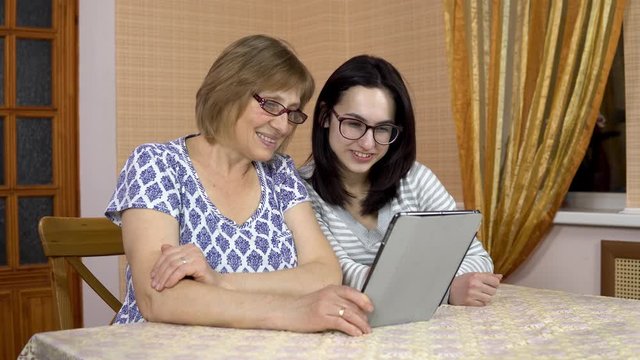 The daughter and the Mother communicate with friends via video link through the tablet. A young woman and an old mother communicate and wave their hands, looking at a tablet. The family is sitting in
