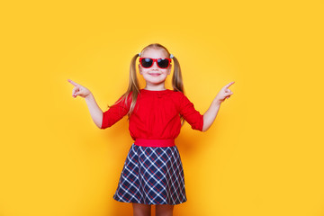 Little girl wearing sunglasses smiling and pointing to the side with thumb up.