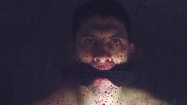 Kidnapped Kid hostage with a tied mouth. Behind a transparent glass covered with bloody drops on a dark background. Ask for help. Close up