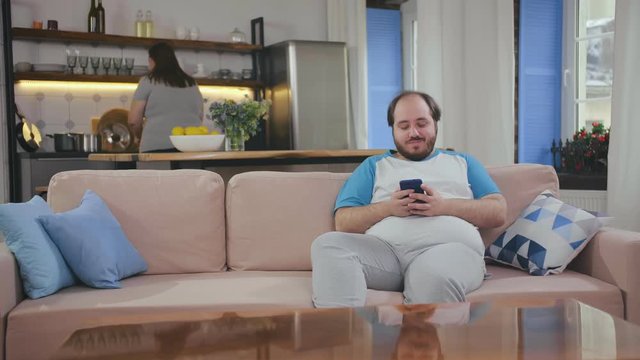 Fat guy relaxing on sofa and chatting with friends on smartphone.