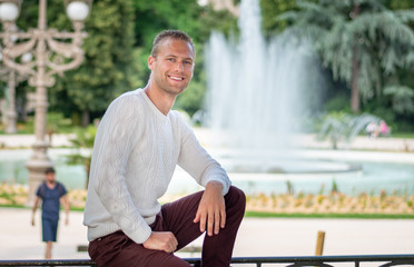 Portrait beau gosse charmant jeune homme caucasian blond aux yeux bleu joyeux et confiance en soi séduisant chanceux charismatique européen heureux zen à motivation pull blanc jardin de la fontaine