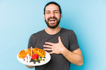 Young caucasian man eating a waffle dessert isolated laughs out loudly keeping hand on chest.