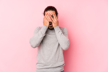 Young long hair man isolated on a pink background blink through fingers frightened and nervous.