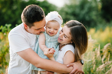 Fototapeta premium The daughter hugging parents on nature. Mom, dad and girl toddler, walk in the grass. Happy young family spending time together, outside, on vacation, outdoors. The concept of family holiday.