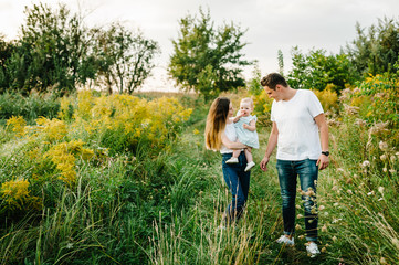Naklejka na ściany i meble The daughter hugging parents on nature. Mom, dad and girl toddler, walk in the grass. Happy young family spending time together, outside, on vacation, outdoors. The concept of family holiday.
