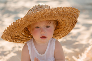 baby girl in white clothes and a straw hat sits on the white sand on the beach in summer