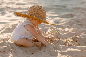 baby girl in white clothes and a straw hat sits on the white sand on the beach in summer