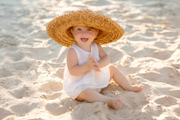 baby girl in white clothes and a straw hat sits on the white sand on the beach in summer