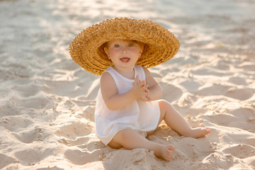 baby girl in white clothes and a straw hat sits on the white sand on the beach in summer