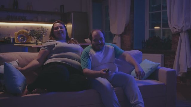 Overweight lazy couple watching tv at home in evening