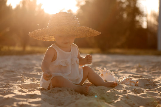baby girl in white clothes and a straw hat sits on the white sand on the beach in summer