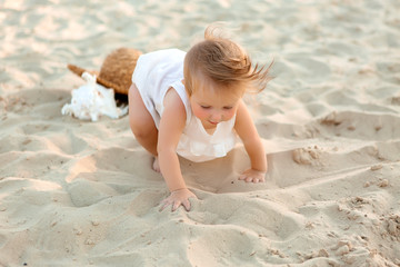 baby girl in white clothes and a straw hat sits on the white sand on the beach in summer