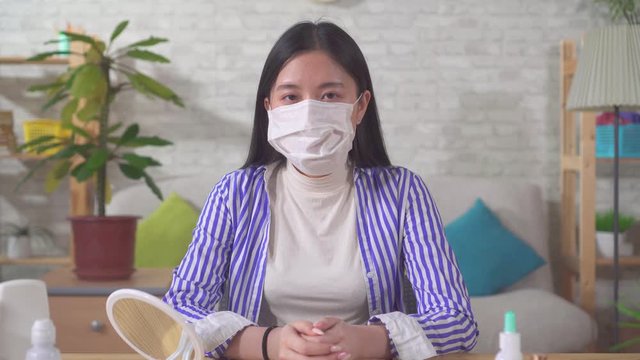 Young Asian Woman Puts On A Medical Mask While Sitting At Home At A Table And Looks At The Camera