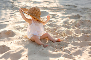 baby girl in white clothes and a straw hat sits on the white sand on the beach in summer