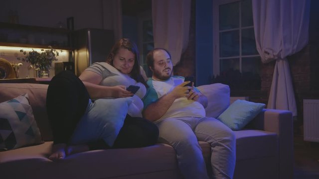 Young fat couple sitting on the sofa at home and connecting online with mobile devices