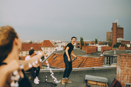 Cheerful man and woman playing with string light on terrace during rooftop party