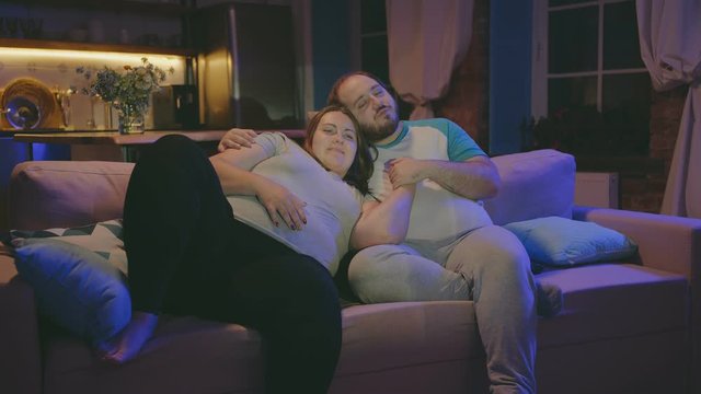 Cheerful obese couple relaxing on couch in evening and watching tv