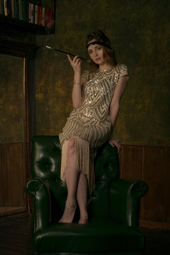 Young adult female model wearing golden pattern vintage style dress and accessories posing on leather armchair holding mouthpiece with smoking cigarette. Roaring twenties noir style concept.