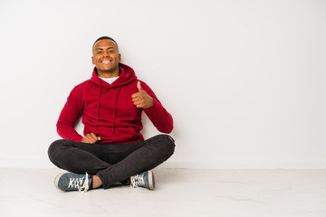 Young latin man sitting on the floor isolated smiling and raising thumb up