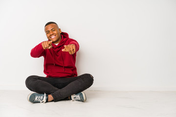 Young latin man sitting on the floor isolated cheerful smiles pointing to front.
