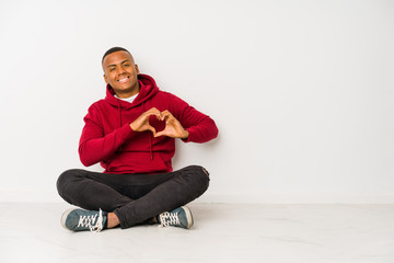 Young latin man sitting on the floor isolated smiling and showing a heart shape with hands.