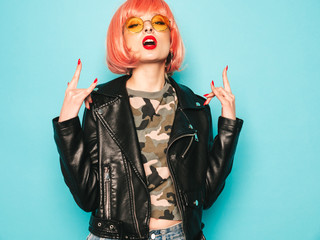Young beautiful hipster bad girl in black leather jacket and earring in nose.Sexy carefree  woman posing in studio in pink wig near blue wall.Confident model in sunglasses.Shows rock and roll sign