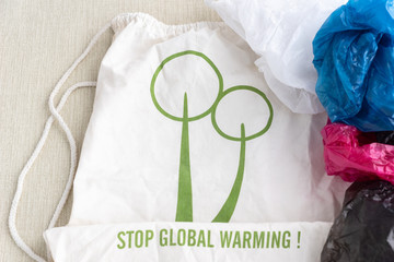 cotton shopping bag with inscription stop global warming on it in center of photo and colorful plastic  or polyethylene bags in right side on light green background,  ecological concept