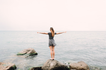 a young girl in a dress with her arms outstretched to the side stands on the sea shore on large stones