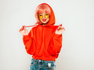 Young beautiful hipster bad girl in trendy red summer hoodie and earring in her nose.Sexy carefree woman in pink wig posing in studio on gray background.Hot model in sunglasses