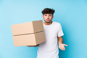 Young arab man holding boxes isolated shrugs shoulders and open eyes confused.