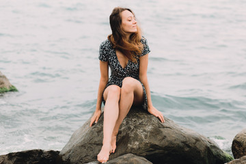 a young beautiful girl in a dress is sitting on large rocks near the sea, the wind is developing her hair