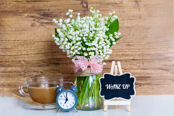 Bouquet of Lily of the Valley  with Alarm Clock,Cup of Coffee and Wake up Text on  Wooden Background .Spring Morning Concept 