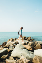 a young girl traveler with a backpack walks barefoot on large stones on the sea coast