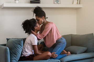 Joyful black young girlfriends hugging and kissing on couch at home