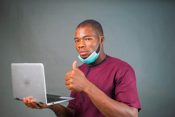 young handsome nigerian man isolated over grey background wearing face mask and using his laptop because of virus outbreak.