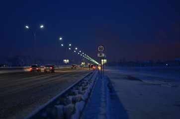 Obraz premium Winter night road at the entrance to the city. The lights of the lampposts and the headlights of cars. It's snowing. Selective focus. A blur of lights.