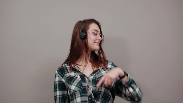 Young brunette girl blue green in checked shirt on grey background happy woman in glasses listens to music in headphones, happily dancing moving her hands