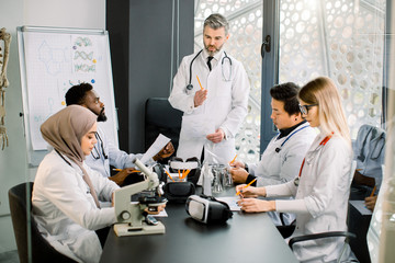 Multi ethnic four young doctors and their teacher professor having a discussion during the conference with documents at the table in the modern office room. Medicine, science, biochemistry concept